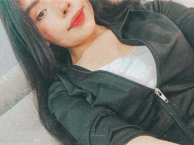 AliceEvansLove with long black hair, smiling, wearing a green zip-up jacket over a white tee, her cute face and small tits visible in the selfie.