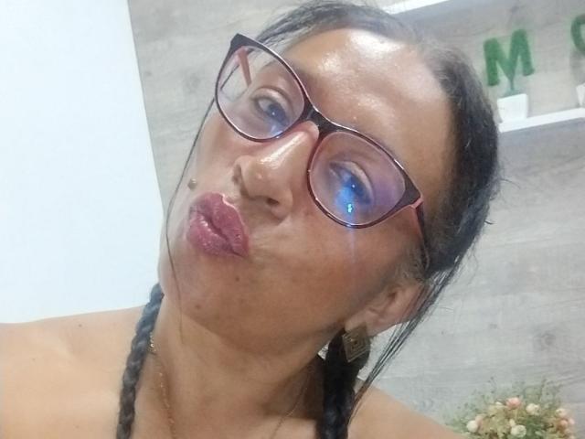 SoffieMature with puffy red lips, wearing glasses, making a pouty face, has black braided hair, and shiny skin. Her tits are not visible in this shot.