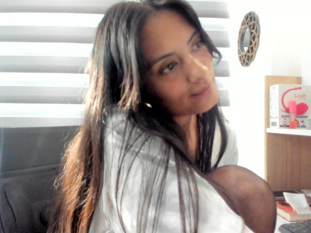 NiaWhitee with long black hair, sitting, wearing a white shirt, looking at the camera, slightly smiling, with her large tits partially visible, in a softly lit room.