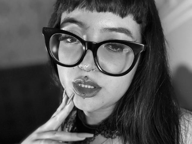 CherryMeloon has black cat-eye glasses, dark lipstick, a septum ring, a lip ring, and a nose ring, with dark straight hair and a hand near her chin.