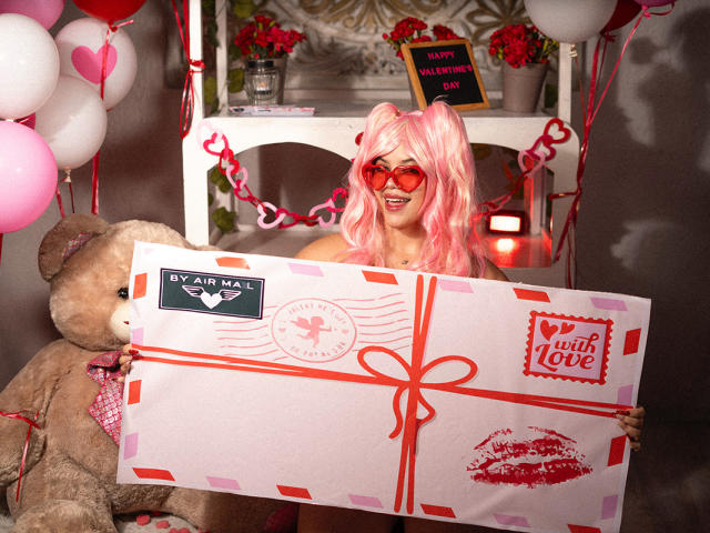 AryButty con i capelli rosa, occhiali da sole a forma di cuore rossi, che tiene in mano una scatola regalo bianca con un nastro rosso, in piedi di fronte a un grande orsacchiotto, allestimento per San Valentino..