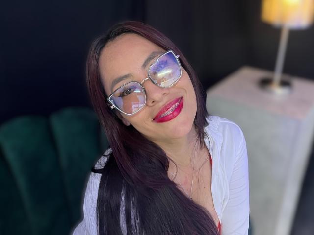 SiennaMoretti with long dark brown hair, red lipstick, and glasses, smiling at the camera, wearing a white top, showing her boobs slightly in the frame.
