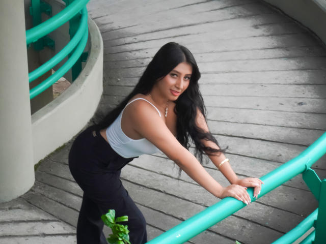 Catalina Rivero, avec ses longs cheveux noirs, son débardeur blanc et son pantalon noir, appuyée sur des rampes vertes, souriant, montrant sa petite poitrine et ses jolies fesses, sur une terrasse en bois..