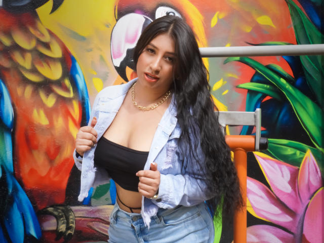 Catalina Rivero, avec ses longs cheveux noirs, portant une veste en jean blanc, un haut noir moulant et un jean bleu, mettant en valeur sa poitrine généreuse, un collier en or et une expression espiègle..
