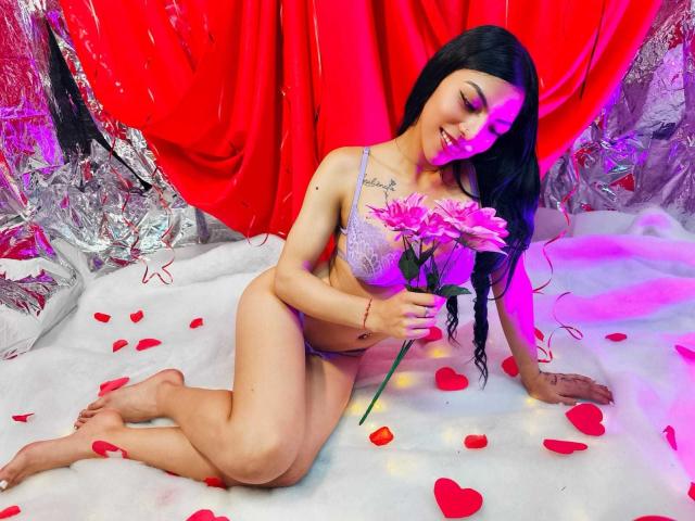 RoxannaMoretti, with long black hair, wears lavender lace lingerie, holding a pink flower, kneeling on white fluffy ground with red petals, under purple and pink lighting.