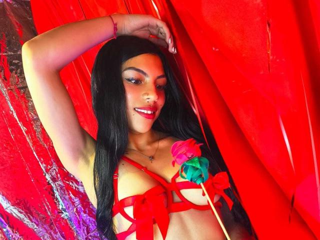 RoxannaMoretti with long black hair, red strappy lingerie, holding a pink rose, arm raised, smiling, red and shiny background, wearing red bracelet, pink lipstick.
