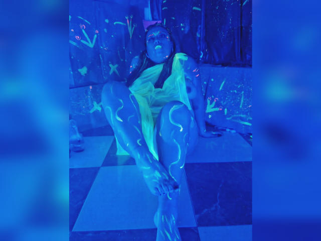 LexyDom is sitting with legs spread, glowing blue liquid covering her shiny skin, yellow dress, and dark hair. Her tits are visible, and her pussy is slightly obscured.