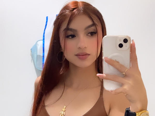 MilanMoon with long brown hair, wearing a brown top, taking a selfie with a white phone, showing her tits and gold necklace, wearing gold hoop earrings and rings.