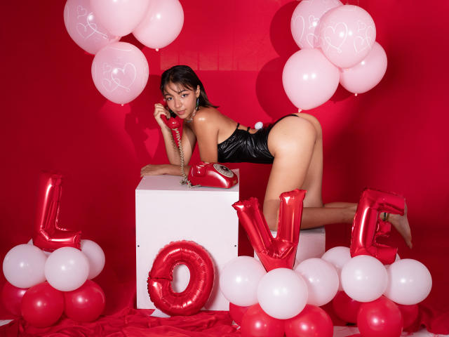 BellaFerrer with black latex outfit, leaning on white cube, holding red heart balloon, surrounded by red and white balloons, red "LOVE" letters, and red background.
