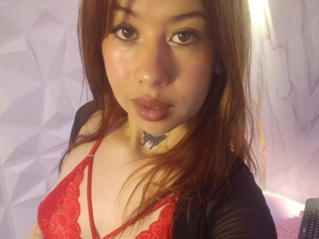 AriSmith with light brown skin, red lace bra, black cardigan, and butterfly tattoo on neck, looking at camera with pouty lips. Red-brown hair, brown eyes, subtle makeup.