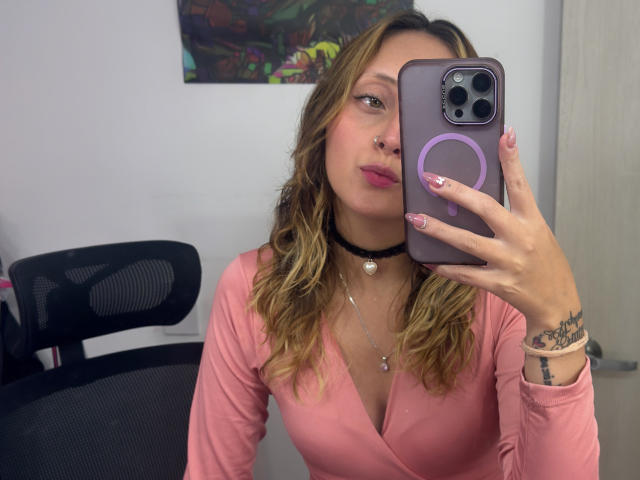 SophiyeGomez with wavy blonde hair, pink top, black choker, nose ring, and pink nails, taking a selfie with a purple phone, showing her tits and tattoos.