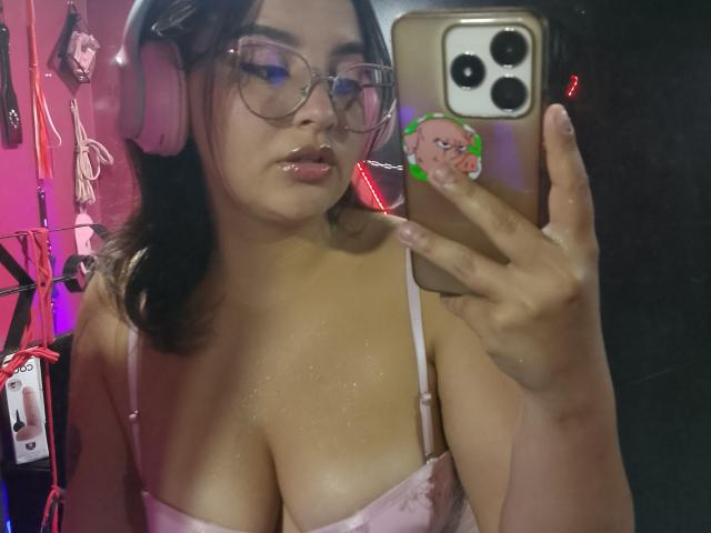 MissSubmissive mit Brille, rosa Kopfhörern und einem pinken Spitzen-BH, die ein Handy mit einem niedlichen Gesicht-Sticker hält und in einem Spiegel-Selfie ihre glänzenden, prallen Brüste zeigt..