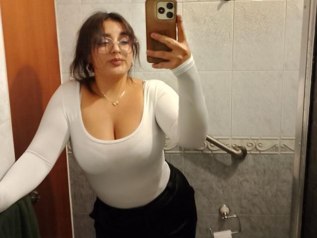 MissSubmissive mit Brille, weißem Langarmshirt und schwarzem Rock, die in einem Spiegel-Selfie im Badezimmer ihre Brüste zeigt. Sie hält ein Telefon, hat braunes Haar, eine Halskette und lächelt leicht..