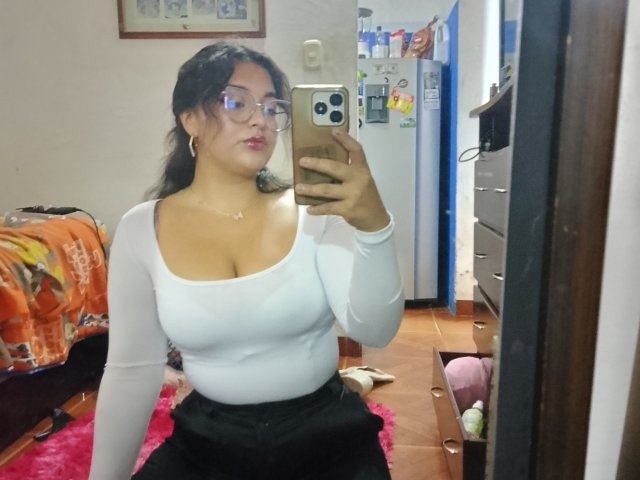 MissSubmissive mit großen Brüsten und dunklem Hintern, trägt eine Hose, eine Brille und ein weißes Oberteil mit langen Ärmeln und macht ein Selfie vor einem Spiegel in einem Schlafzimmer. Sie hält ein goldenes Handy..