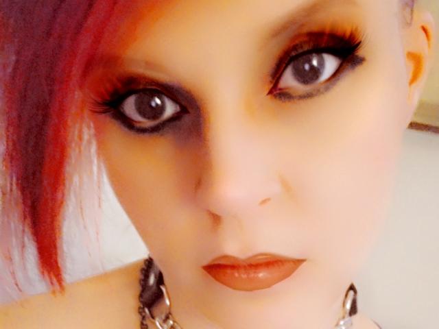 UnLaced has red hair, dark eye makeup, and red lipstick. She's looking up with large, dark eyes, wearing a metal chain necklace. Her tits are not visible.