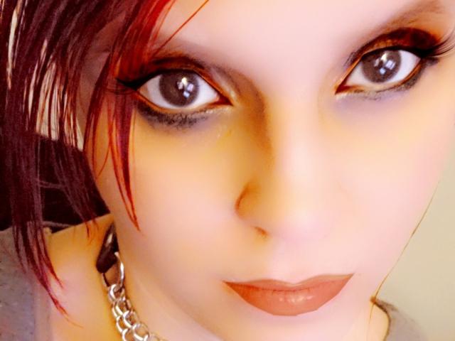 UnLaced has red hair, pale skin, and dark, dramatic eyeliner. She's wearing a silver chain necklace and has red lipstick. Her wide eyes are looking directly at the camera.