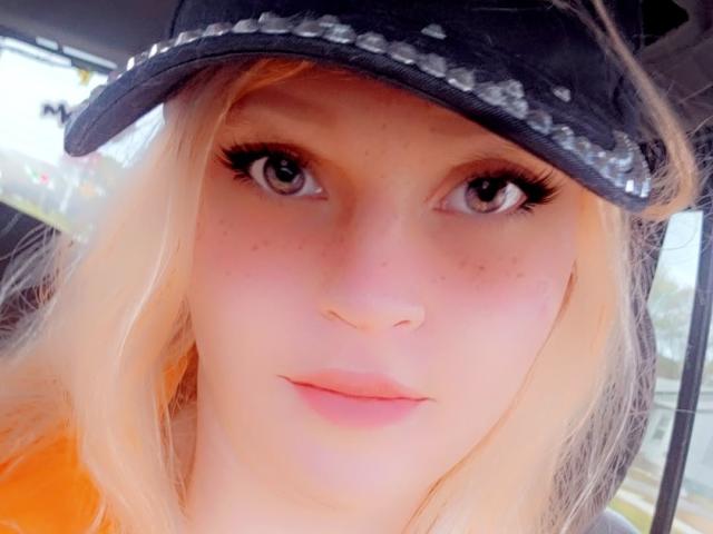UnLaced has blonde hair, blue eyes, and a cute face with a slight smile, wearing a black hat with silver studs. She's freckled and has long, thick eyelashes.