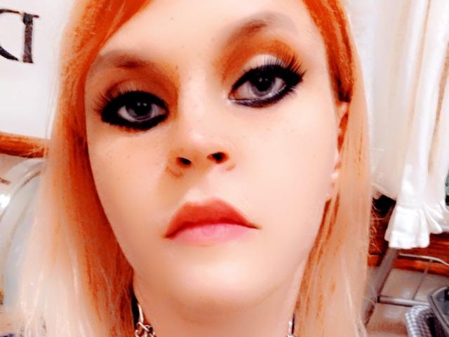 UnLaced has orange hair, black eyeliner, and full lips, with a nose piercing and a serious expression, wearing a choker. Her eyes are wide with thick, black lashes.