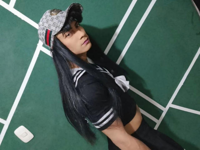 Sofia69xx with long black hair, wearing a sailor outfit, baseball cap with red stripe, looking over shoulder, green carpet, white lines, showing black shorts, and pink lipstick.