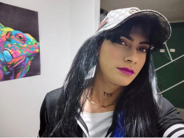 Sofia69xx with long black hair, purple lipstick, and a black-and-white cap, wearing a black jacket over a white shirt, has a small tattoo below her neck.