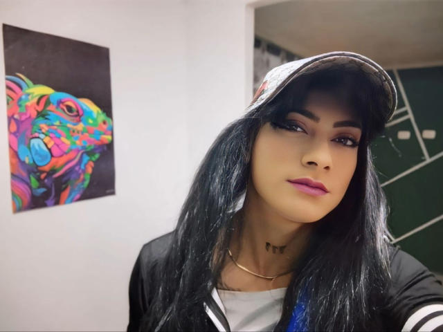 Sofia69xx with long black hair, purple lipstick, wearing a cap and black shirt, has a necklace, and is looking at the camera, with colorful art in the background.