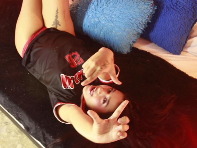 MissAris is lying on a black surface, wearing a black sports jersey with red and white trim, showing off her tits and pointing at the camera with her fingers.