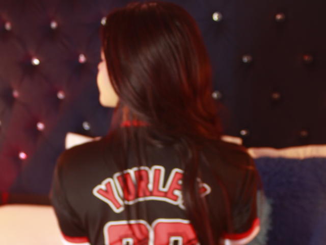 MissAris with long black hair, back to camera, wearing a black and red football jersey with "JUDSON 98" on it, sitting on a bed with blue and white pillows.
