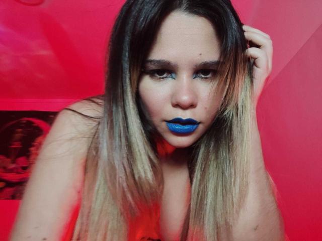 AshGraven with blue lipstick, blonde-to-brown ombre hair, and serious eyes, hand in hair, red-lit background, looking at camera, upper body, slightly tilted.