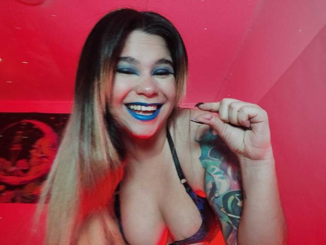 AshGraven with blue lipstick, smiling, pointing with black-nailed finger, long blonde hair, tattooed arm, cleavage in black top, red lighting, and confident expression.