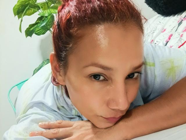 AmberDemons with red hair, lying down, head on white pillow, wearing pink earrings, looking at camera with slight pout, arms folded, wearing light-colored shirt, green plant in background.