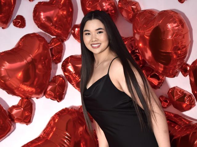 AmandaPresle with long black hair, smiling, wearing a black satin dress, standing in front of red heart-shaped balloons. She's cute with a small ass and slim build.