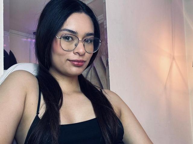 IsaDoux with dark long hair, glasses, black tank top, showing her tits, sitting in a chair, slightly smiling, pink lipstick, white wall in background, soft lighting.