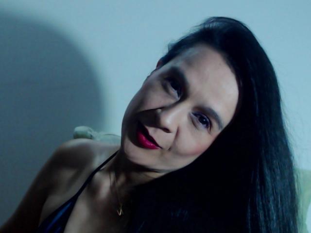 AileenRamirezW with dark hair, red lipstick, and a black halter top, leaning forward with a slight smile, shadow on the left side of her face.