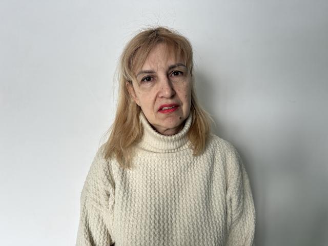 PattySanny has shoulder-length blonde hair with a slight part. She wears a cream-colored, knitted turtleneck sweater with a textured, ribbed pattern. Her skin is fair with visible wrinkles and small freckles on her cheeks. She has red lipstick on and looks directly at the camera with a slight, neutral expression. The image is centered on PattySanny, who is facing forward with a simple, white background. The lighting is even, highlighting the texture of her sweater and the subtle details on her face.