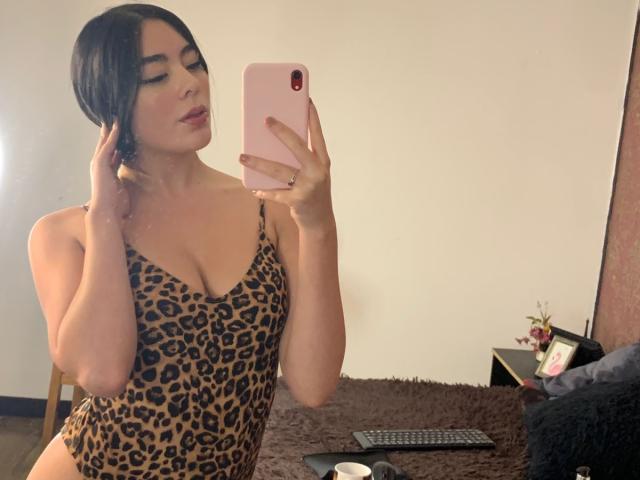 Samantha Chasse mit dunklem Haar, die ein Leopardenmuster-Dessous trägt, hält ein rosa Telefon, macht ein Selfie im Spiegel und zeigt ihre Brüste und ein leichtes Lächeln, im Schlafzimmer..