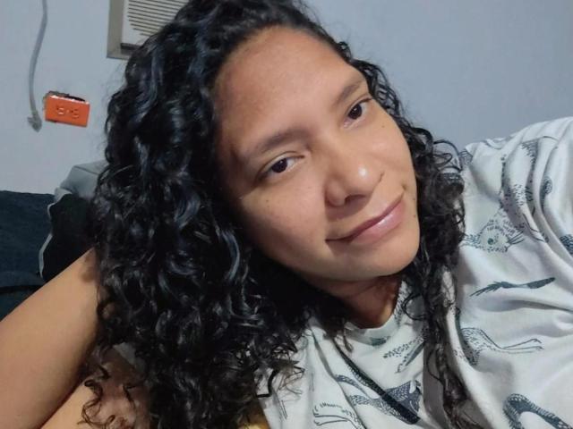 GabiGrandeth has curly black hair, a light brown skin tone, and is smiling with a small nose, wearing a white shirt with black prints, in a casual selfie.