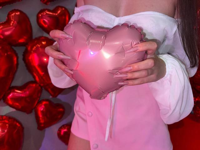 Rouzi holding a shiny pink heart-shaped balloon with both hands, wearing a white off-shoulder shirt and pink button-up, with long, pink-tinted nails. Red heart balloons in background.