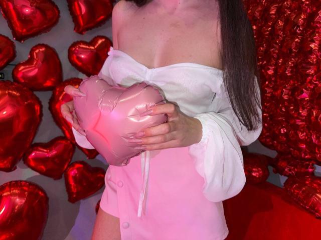 Rouzi tiene in mano un palloncino rosa a forma di cuore, indossa una maglietta bianca con una spalla scoperta, che mette in mostra il suo seno, ha lunghi capelli castani, una pelle leggermente rosata, ed è sullo sfondo di un cuore rosso..