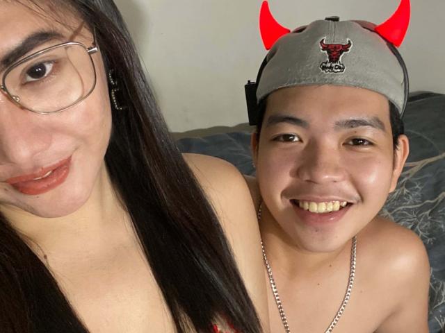 A selfie photograph of AsianCoupleDesire. AsianCoupleDesire on the left has long black hair, glasses, and light brown lipstick, smiling slightly. AsianCoupleDesire on the right, shirtless, wears a gray baseball cap with red devil horns and a red bull emblem, smiling widely. Both have light brown skin. The right AsianCoupleDesire has a necklace and a slightly chubby face. The left AsianCoupleDesire's face is partially in the frame. The right AsianCoupleDesire's cap is slightly worn. The image has a casual, intimate feel.