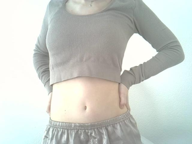 A close-up photograph of DitaVira from the chest to the waist. She wears a ribbed, long-sleeve, cropped, beige top that reveals her midriff and a pair of loose, beige pants with an elastic waistband. Her right hand is on her hip, slightly pushing the top up. Her skin is light, and she has a small, visible navel. The image is brightly lit, emphasizing the texture of the top and the smoothness of her skin.