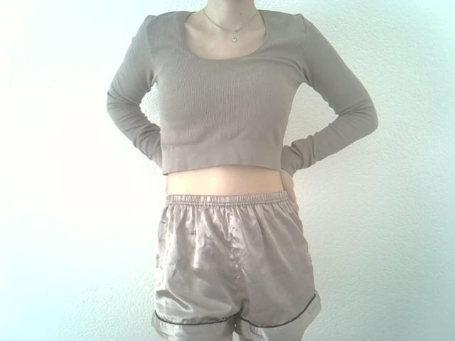 DitaVira stands with hands on hips, wearing a beige rib-knit long-sleeve crop top and matching satin shorts. Her top is slightly tight, revealing a small section of her midriff. The shorts are loose, with a satin sheen and black trim. Her neck has a delicate silver necklace. The composition is simple, with a white textured wall in the background. The image focuses on her upper body and shorts.