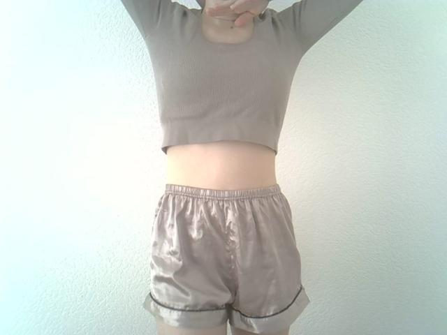 DitaVira stands with arms raised, wearing a light gray, long-sleeve crop top and matching high-waisted shorts. The shorts have a satin-like texture, slightly shiny, and are rolled up at the bottom. The crop top is slightly cropped, revealing a small portion of her midriff. The image is well-lit with soft, even lighting, highlighting the smooth texture of the shorts and the casual, relaxed style of the outfit. The focus is on her torso and the upper part of her body.