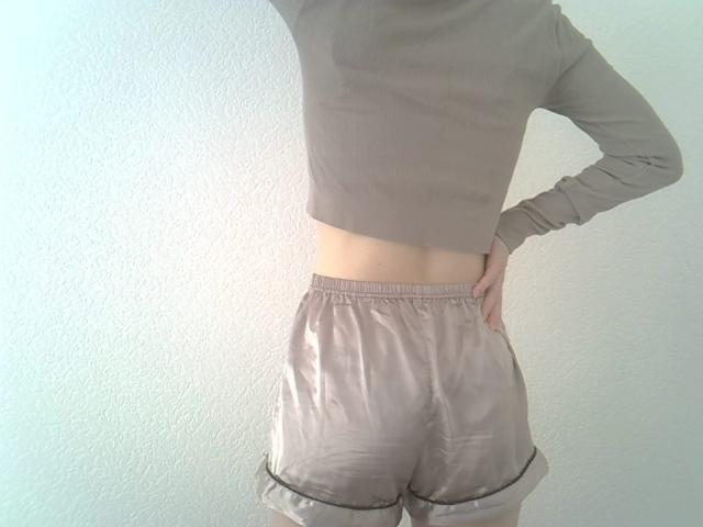 DitaVira stands with her back to the viewer, right hand on her hip. She wears a long-sleeve, cropped beige top and matching silky shorts with a black trim at the bottom. Her skin is light, and her midsection is exposed. The shorts have a smooth, shiny texture, contrasting with the matte top. The image focuses on her lower back and shorts, with her head out of the frame. The background is a plain, textured wall.
