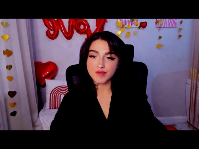 RaspberryGirl with dark hair, full lips, and subtle makeup, wearing a black top, sitting in a chair, surrounded by red and yellow heart decorations.