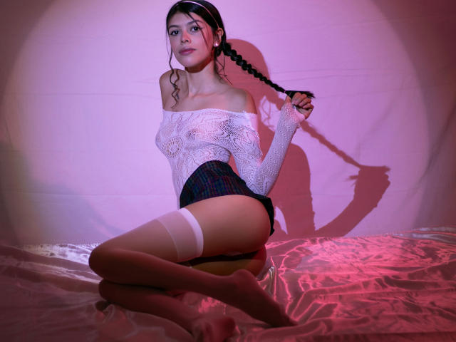 KeityRousse with braided hair, white lace off-shoulder top, plaid skirt, and white thigh-highs, kneeling on shiny pink bed, holding her braid, with a sexy, confident expression.
