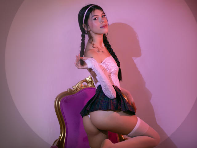 KeityRousse with braided black hair, white off-shoulder top, plaid skirt, white thigh-highs, showing her ass, kneeling on a purple velvet chair, under pink and purple lighting.