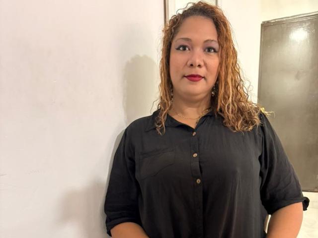 IvyQueen has curly brown hair, red lipstick, and is wearing a black button-up shirt. She's standing against a white wall with a metal door in the background.
