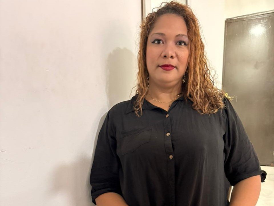 IvyQueen has curly brown hair, red lipstick, and is wearing a black button-up shirt. She's standing against a white wall with a metal door in the background.