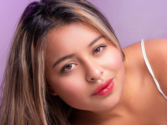 AngellVisen has tan skin, brown eyes, and long blonde hair with darker roots. She's wearing pink lipstick and a white strap, smiling slightly with her head tilted.