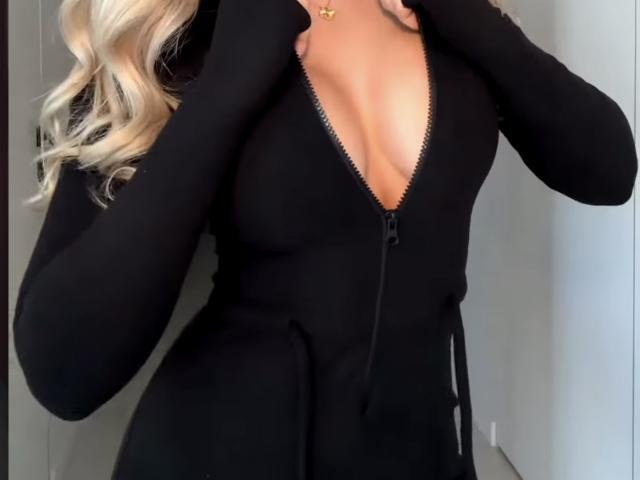 MargoFun, com cabelo loiro, vestindo um vestido preto com zíper, mãos no peito, decote profundo, zíper preto, mangas pretas, colar dourado, luz suave, interior, close-up..